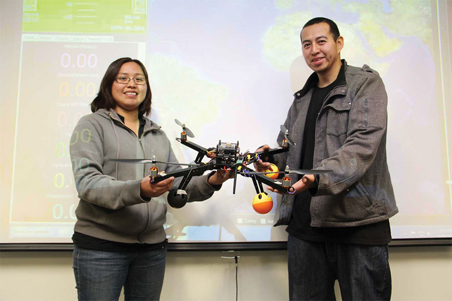 Two individuals stand in front of a screen while holding a drone