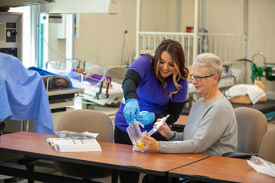 A respiratory therapy student assists an older woman with a breathing exercise using a medical training device in a clinical classroom. The image highlights hands-on learning in respiratory therapy and offers a glimpse into what respiratory therapists do in their careers.