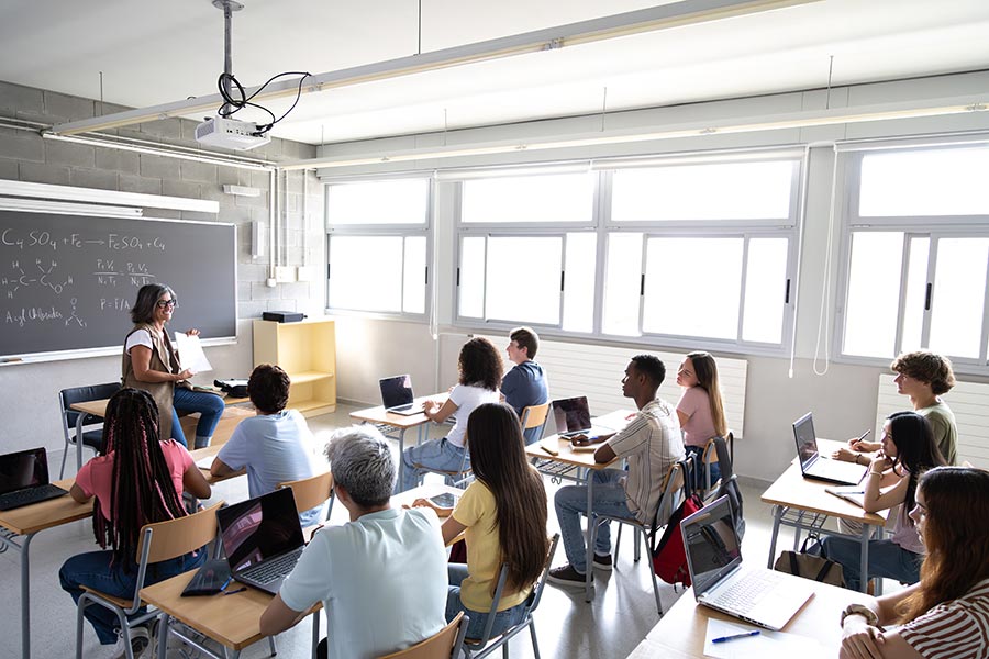A diverse group of high school students sits at desks with laptops, attentively listening to a certified teacher standing at the front of a bright, modern classroom. This image represents a typical teaching environment in New Mexico and supports those exploring how to become a teacher, how to become a certified teacher, or how to change career to teaching.