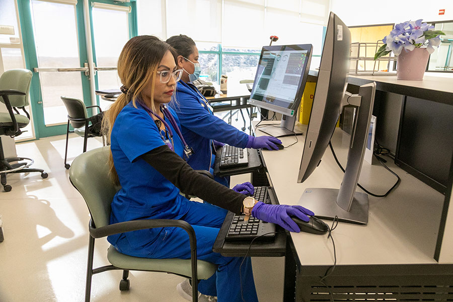 Certified Nursing Assistants (CNAs) in blue scrubs and gloves work at computer stations in a clinical setting, illustrating the administrative and patient care duties CNAs perform daily