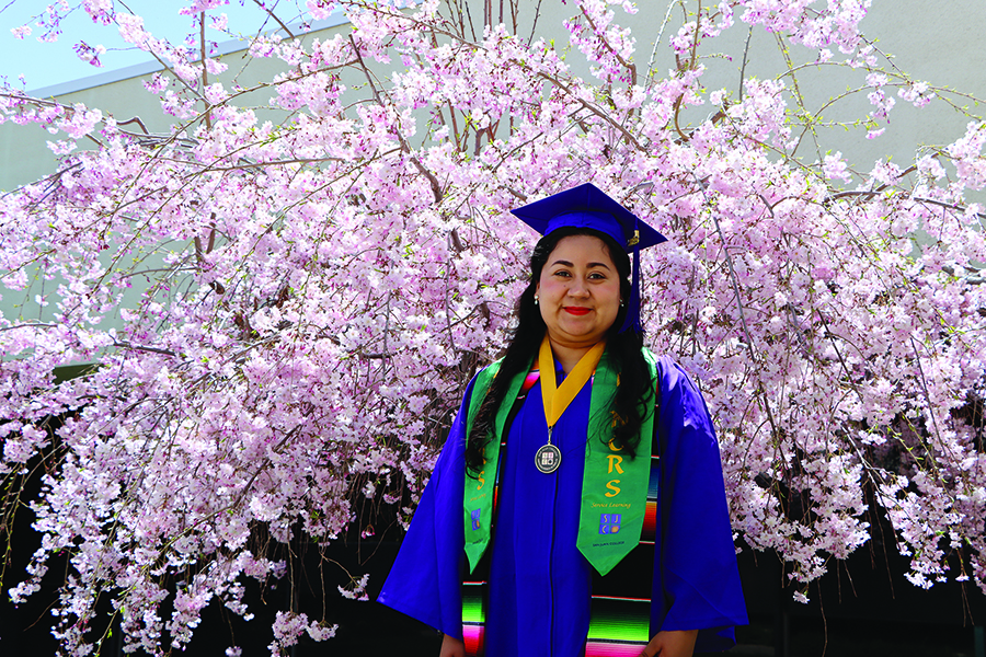 Graduate in full gown with honors tassels in front of pink flowering tree