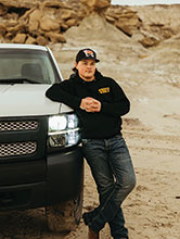 Talon Sheek leaning against his white truck in a dirt area with large rocks in background