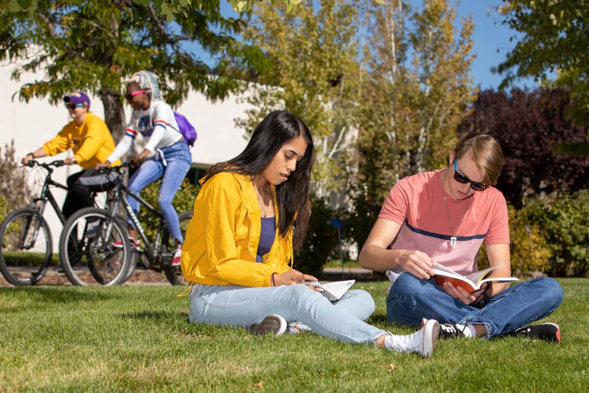 Two students riding bikes and two students studying while sitting on lawn.
