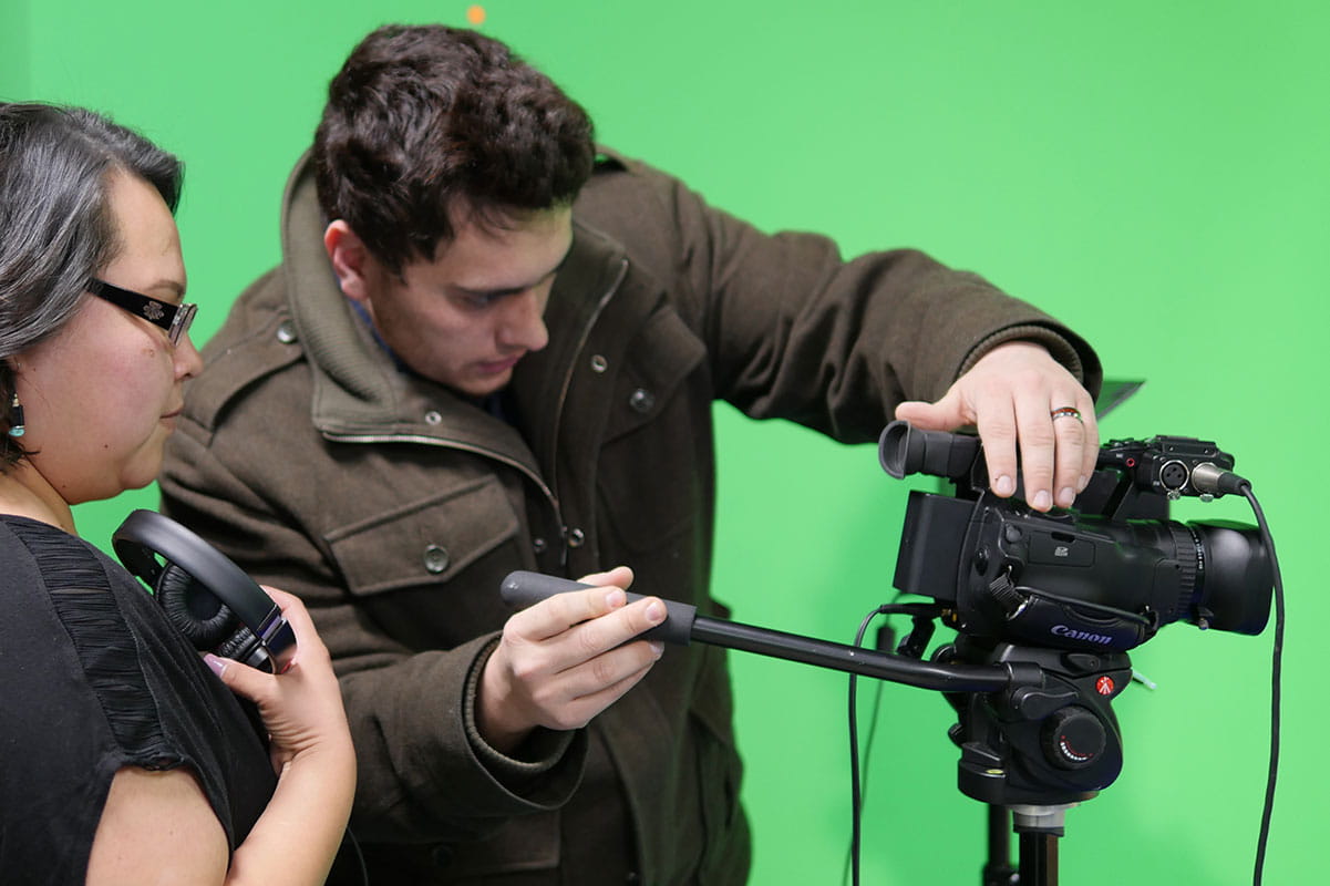 Two individuals work with a camera in front of a green screen
