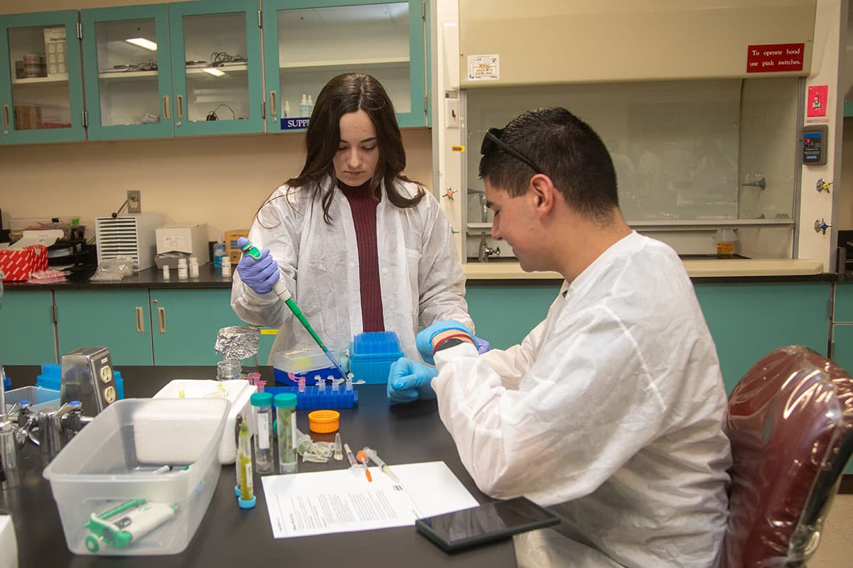 Two students in lab coats work with pipettes and test tubes at a table in a lab classroom