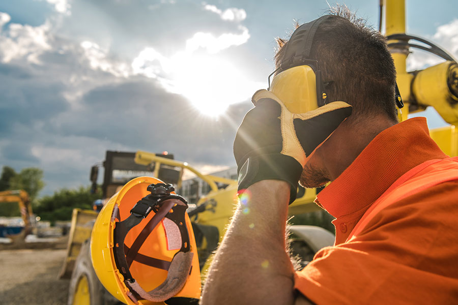 A construction worker takes off a helmet and ear protectors with construction equipment visible in the background
