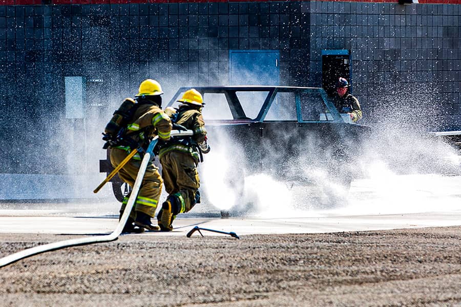Two firefighters drag a water hose toward a burning car