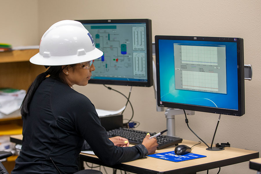 An individual in a white hard hat and safety goggles sits at a desk looking at a computer screen
