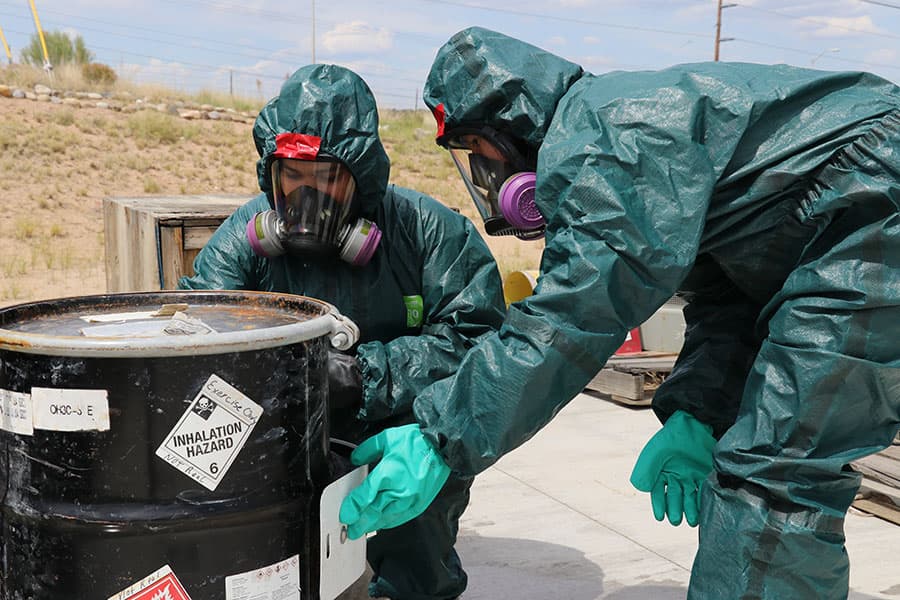 Two individuals in hazmat suits inspect a container of hazardous materials