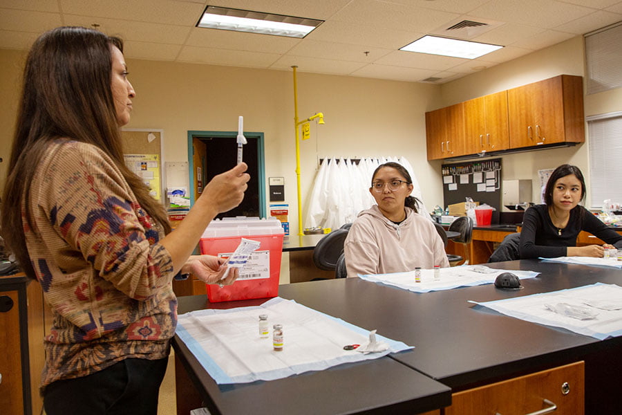 An instructor holds a medical syringe while standing in front of a table while two students look on