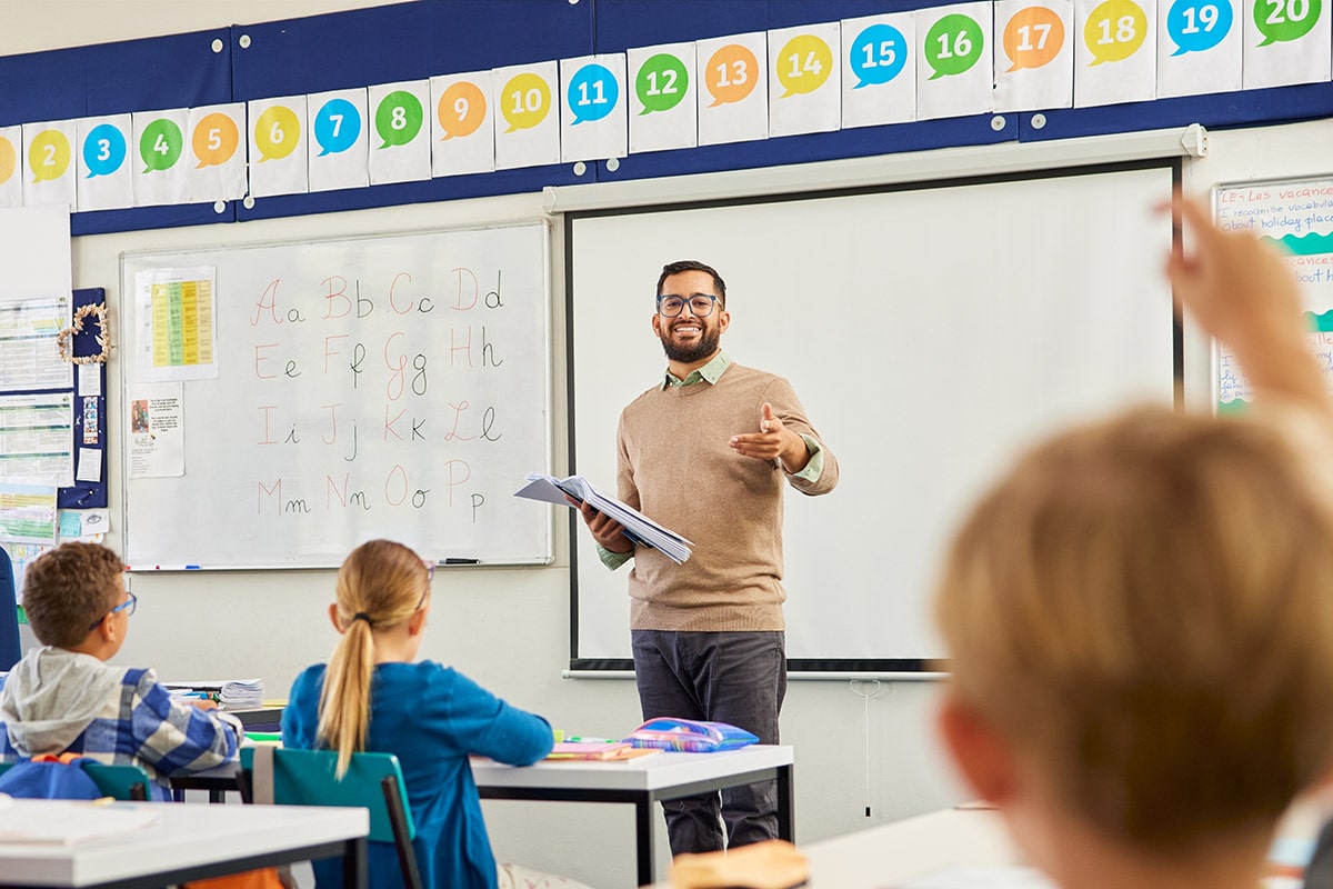 Man standing at the front of a classroom pointing at a student with their hand up to answer a question.