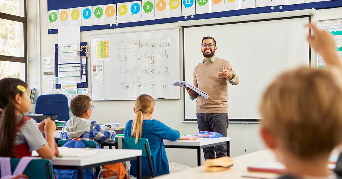Man standing at the front of a classroom pointing at a student with their hand up to answer a question.