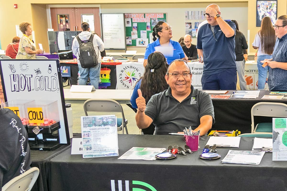 A man smiles a gives a thumbs-up at one of the many employer tables during a community job fair on campus.